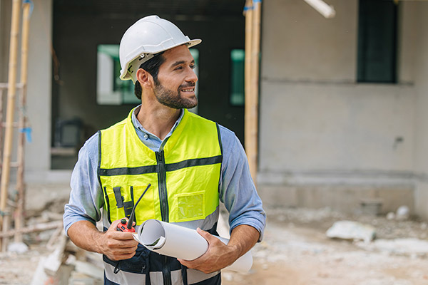 Smiling construction engineer wearing a safety vest and hard hat holding blueprints and a two-way radio at an active job site.