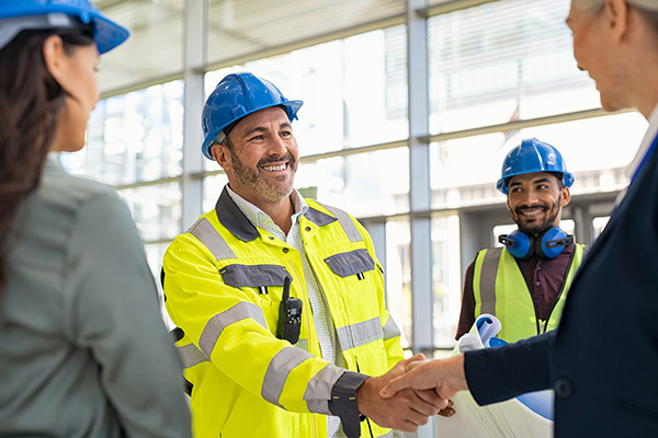 Construction site meeting showing a smiling worker in a yellow safety jacket and blue hard hat shaking hands with a business professional while colleagues look on.