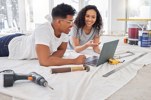 Young couple lay on the floor of their renovated home, looking at a computer.