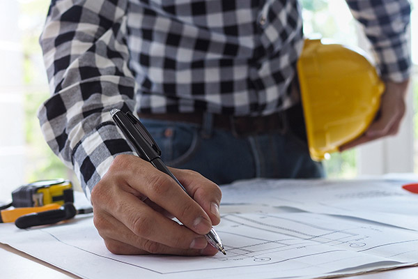 A contractor's handwriting on blue prints while the other holds a hard hat on his hip.
