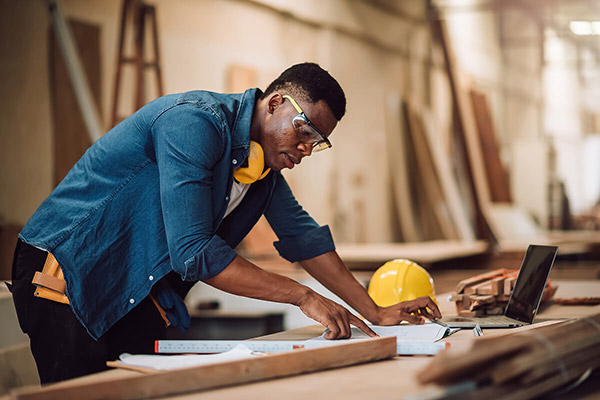 A contractor with safety glasses reviews documents and a laptop at a workbench in a workshop environment.