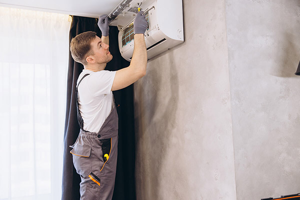 A technician in overalls and gloves works on installing or repairing a white air conditioning unit on a wall